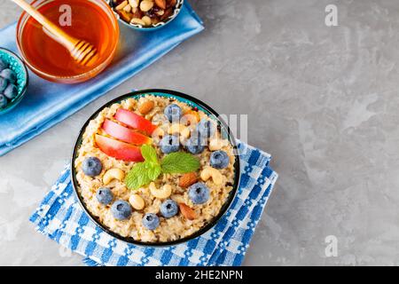 Haferflocken und die Zutaten für ein gesundes Frühstück auf grauem Hintergrund. Gekochte Haferflocken, Heidelbeeren, Nüsse, Apfelscheiben und Honig. Gesundes Frühstück Stockfoto