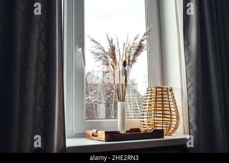 Mit getrockneten natürlichen verschiedenen Graspflanzen innerhalb Vase drinnen für die Heimtextilien. Vase mit verschiedenen Pflanzen und Holz schwarz Metalldraht Laterne auf Fenster. Stockfoto