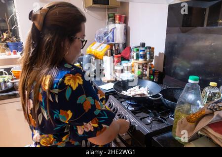 Eine Frau rührt Huhn in einem Wok auf einem Gasherd in einem britischen Haus, umgeben von Alltagsgewirr. Stockfoto