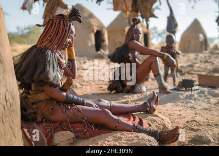 Himba-Frauen sitzen vor ihren Hütten in einem traditionellen Himba-Dorf in der Nähe von Kamanjab im Norden Namibias, Afrika. Stockfoto