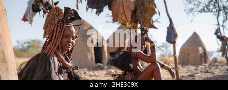 Panoramaaufnahme mit Himba-Frauen, die vor ihren Hütten in einem traditionellen Himba-Dorf in der Nähe von Kamanjab im Norden Namibias, Afrika, sitzen. Stockfoto