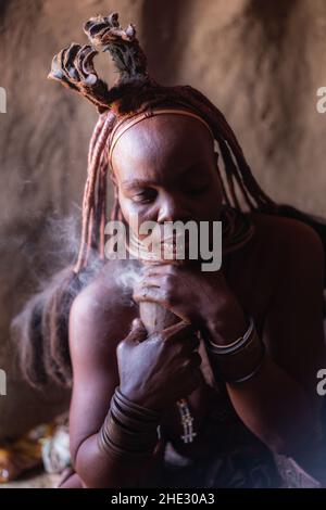 Himba-Frau nimmt täglich ein Rauchbad in ihrer Hütte in einem traditionellen Himba-Dorf in der Nähe von Kamanjab, im Norden Namibias, Afrika. Stockfoto