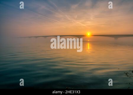 Lange Brücke über glasig glattes Wasser geht bis in die Unendlichkeit über den herrlichen Ozean unter dem wunderschönen bunten Himmel in Zeeland, den Niederlanden Stockfoto