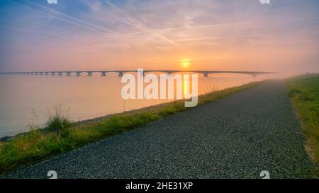 Lange Brücke über glasig glattes Wasser geht bis in die Unendlichkeit über den herrlichen Ozean unter dem wunderschönen bunten Himmel in Zeeland, den Niederlanden Stockfoto