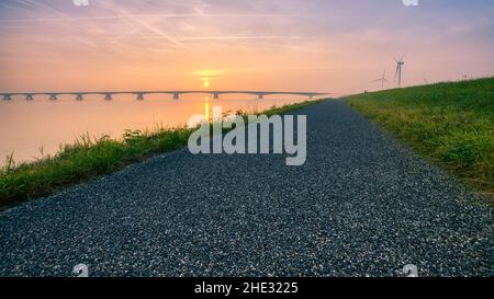 Lange Brücke über glasig glattes Wasser geht bis in die Unendlichkeit über den herrlichen Ozean unter dem wunderschönen bunten Himmel in Zeeland, den Niederlanden Stockfoto