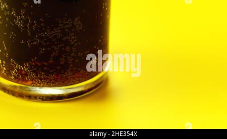 Glas mit Cola auf gelbem Tisch, Nahaufnahme von Blasen in brauner Flüssigkeit Stockfoto