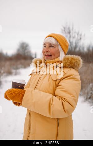 Frau in gelber Winterkleidung hält heißen Kaffee oder Tee aus der Papiertasse draußen im Winterpark Stockfoto