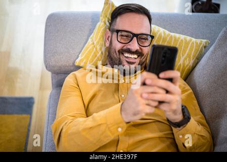 Fröhlicher junger Erwachsener reifer Mann glücklich lächelnd mit Smartphone liegend auf Sofa Couch zu Hause Lesung Nachrichten auf Handy Guy mit Auge Stockfoto