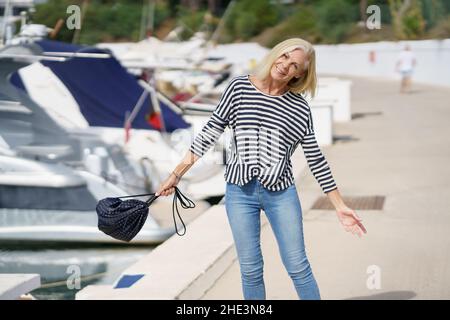 Glückliches Weibchen mit 60s Jahren, das an einem Strandort in der Nähe des Strandes entlang schlendert. Stockfoto