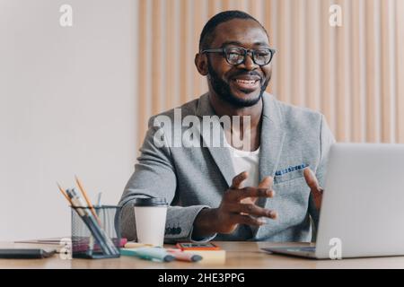 Fröhlicher afroamerikanischer Geschäftsmann mit Brille und stilvollem Blazer, der Videoanruf auf dem Laptop macht Stockfoto