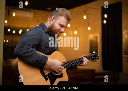 Ein junger Mann mit Bart spielt in einem Raum mit warmer Beleuchtung eine akustische Gitarre Stockfoto