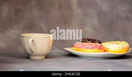Eine Tasse mit einem heißen Getränk und ein Teller mit zwei frischen Donuts in Glasur auf einem grauen abstrakten Hintergrund. Nahaufnahme. Stockfoto