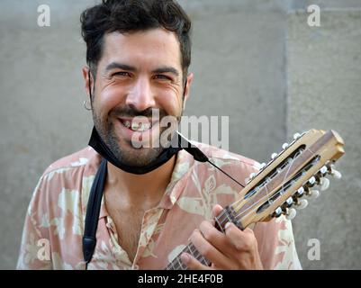 Ein hübscher junger Lateinamerikaner mit schwarzem Bart und Gesichtsmaske unter dem Kinn spielt eine kleine Andengitarre (Charango) mit zehn Saiten. Stockfoto