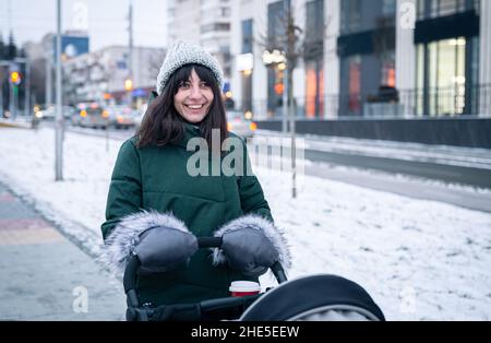 Stilvolle junge Mutter mit einer Tasse Kaffee auf einem Spaziergang mit einem Kinderwagen im Winter. Stockfoto