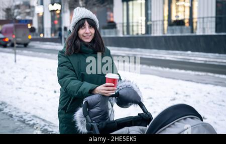 Stilvolle junge Mutter mit einer Tasse Kaffee auf einem Spaziergang mit einem Kinderwagen im Winter. Stockfoto