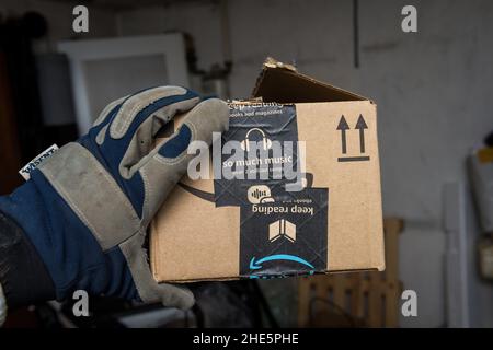 Frankfurt, Deutschland - 10. Dez 2021: Männliche Hand im Schutzhandschuh, der im Lager Amazon hält, öffnet eine Kartons-Box mit weihnachtlichen Logos während der jährlichen E-Commerce-Weihnachtszeit Stockfoto