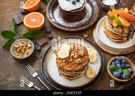 Wir feiern den Pfannkuchen-Tag, ein gesundes Frühstück. Köstliche hausgemachte amerikanische Bananen-Pfannkuchen mit Heidelbeeren, Nüssen, Karamell, Schokolade, roter Orange auf rus Stockfoto