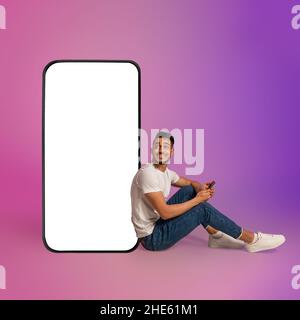 Junger Araber, der Handy benutzt, während er in der Nähe eines riesigen Mobiltelefons mit leerem weißen Bildschirm und Neonlicht sitzt. Modell Stockfoto