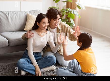 Froh glücklich asiatisch teen Tochter gibt hoch fünf bis junge Eltern sitzen auf dem Boden im Wohnzimmer innen Stockfoto