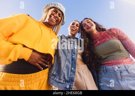 Ansicht von unten von drei Mädchen, die fröhlich lachen. Junge Frauen, die Spaß haben, während sie im Freien in der Stadt zusammen stehen. Gruppe der Generation z frien Stockfoto