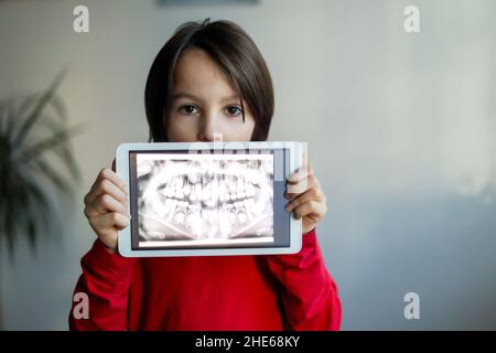 Kind, das Zahnspange trägt, ein Jugendlicher, hält eine Tablette mit einem Bild seiner Röntgenzähne von seinem Gebiss zu Hause Stockfoto