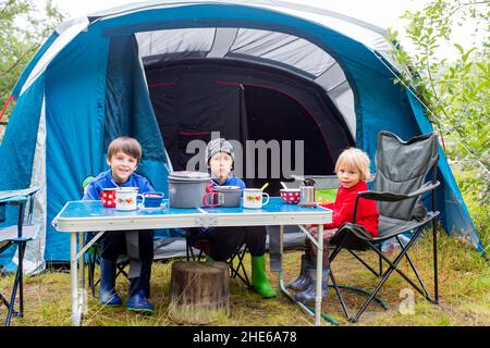 Glückliche Familie, drei Kinder, junge Brüder, Frühstück im Regen während wild Camping in Norwegen, Sommerzeit Stockfoto