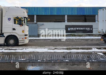 Zwei geparkte Lastwagen. In der Lücke Blick auf ein Werbeplakat einer Wurst-Snackbar. Davor zahlreiche Fahrradständer. Stockfoto