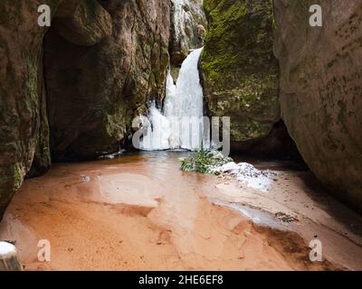 Gefrorener kleiner Adrspach-Wasserfall. Roter Sand, Schnee und Eiszapfen am Bach durch den Wasserfall in den Felsen. Beliebter Felspark an der tschechischen Grenze zu Polen. Stockfoto