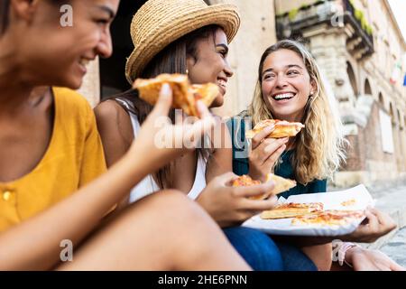 Drei junge, multirassische Frauen lachen beim Essen einer Pizza in Italien Stockfoto