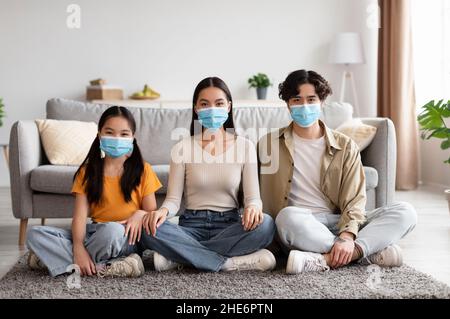 Fröhliches Teenager-Mädchen und junge chinesische Eltern in Schutzmasken sitzen auf dem Boden im Wohnzimmer Innenraum Stockfoto