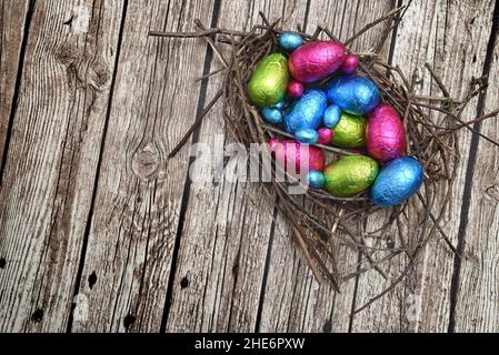 Folienumwickelte bunte ostereier in rosa, grün, blau und gelb in einem natürlichen Nest aus Stäben und Zweigen, vor mehrkörnigem braunen Hintergrund. Stockfoto