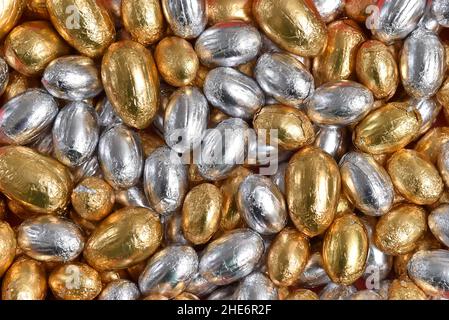 Große und kleine, in Gold und Silber verpackte Ostereier aus Schokolade vor einem rosa, pfirsichfarbenen Hintergrund. Stockfoto