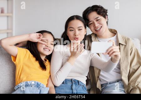 Fröhlich junge asiatische Kerl umarmt Frau, Dame macht weht Kuss, teen Tochter macht Frieden Zeichen und Foto zu machen Stockfoto