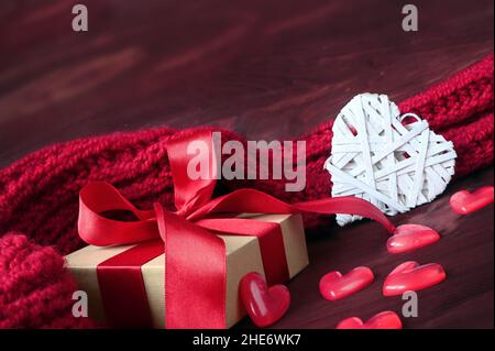 Weiße Geschenkbox und großes Herz mit rotem Strickschal auf rotem Holzhintergrund. Urlaubskonzept. Stockfoto