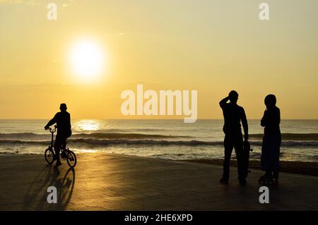 Die Menschen haben sich gegen die Morgensonne auf der gepflasterten Promenade am Strand von Barceloneta in Barcelona, Spanien, geschildet. Stockfoto