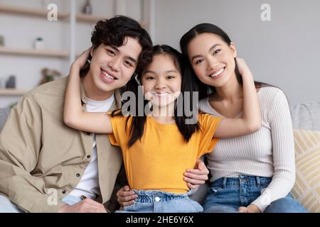 Lächelndes Teenager-Kind umarmt junge asiatische Mama und Papa sitzt auf dem Sofa und schaut in die Kamera Stockfoto