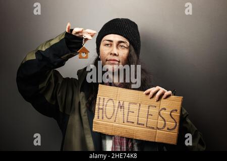 Eine lächelnde Bettlerin hält ein Pappschild mit der Aufschrift Obdachlos und schaut auf einen Schlüsselanhänger in Form eines Hauses. Das Konzept der Unterstützung von VAG Stockfoto