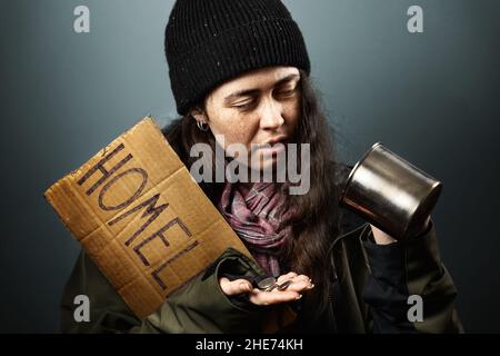 Eine traurige Bettlerin hält ein Pappschild mit der Aufschrift Obdachlos und schaut sich Münzen aus einem Stahl-Almosenbecher an. Dunkler Hintergrund. Das Konzept der Hilfe Stockfoto