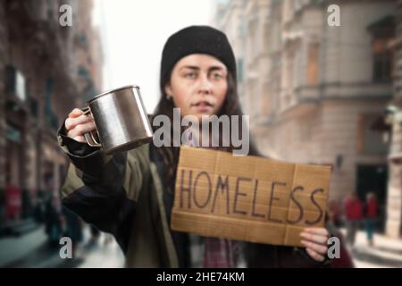 Die unverschämte Bettlerin hält ein Pappschild mit dem Text Obdachlos und einem Almosenbecher aus Stahl. Unschärfer Hintergrund mit Straße und Menschen. Das Konzept von h Stockfoto