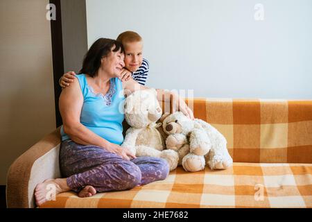 Liebe und familiäre Verbindung. Familienportrait glückliche Großmutter und kleiner Enkel zu Hause. Der Junge, der hinter seiner Großmutter steht, die auf der Couch sitzt, umarmt ihre Schultern neben weichem Spielzeug Stockfoto