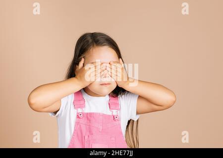 Kind schließt die Augen mit den Händen nicht sehen andere mit geschlossenem Mund nicht reden mit anderen tragen leuchtend rosa Jumpsuit und weißes T-Shirt auf beigem Hintergrund Stockfoto