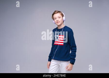 Fröhlicher kaukasischer Junge mit kabelgebundenen Kopfhörern, der Musik mit zufriedenem Gesicht hört. Ein junger Mann genießt seine Freizeit in einem Pullover mit us-Flagge. Innenaufnahme des Studios isoliert auf grauem Hintergrund Stockfoto