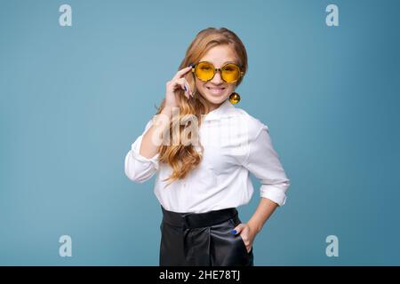 Fröhliche junge Frau mit gelben Gläsern lächelt vor blauem Hintergrund vor der Kamera. Nahaufnahme glücklicher Teenager, der mit einer Brille lacht. Schönes Mädchen, das Spaß mit niedlichen Augen, zeigt großes Grinsen Stockfoto