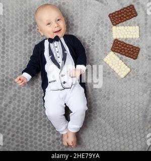 Ein vier Monate altes Baby in einem Smoking liegt an seinem Geburtstag auf einem grauen Teppich mit vier weißen und braunen Pralinen. Lächelnd und launend Baby. Stockfoto