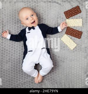 Ein vier Monate altes Baby in einem Smoking liegt an seinem Geburtstag auf einem grauen Teppich mit vier weißen und braunen Pralinen. Lächelnd und launend Baby. Stockfoto