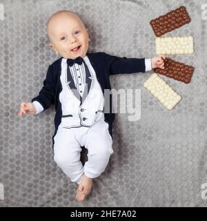 Ein vier Monate altes Baby in einem Smoking liegt an seinem Geburtstag auf einem grauen Teppich mit vier weißen und braunen Pralinen. Lächelnd und launend Baby. Stockfoto