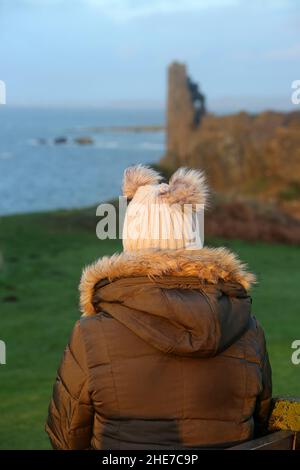 Dunure, Ayrshire, Schottland, Großbritannien. Eine Frau am Wintertag sitzt mit dem Rücken zur Kamera und schaut auf Dunure Castle Stockfoto