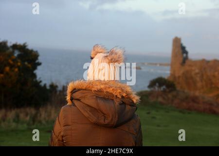 Dunure, Ayrshire, Schottland, Großbritannien. Eine Frau am Wintertag sitzt mit dem Rücken zur Kamera und schaut auf Dunure Castle Stockfoto