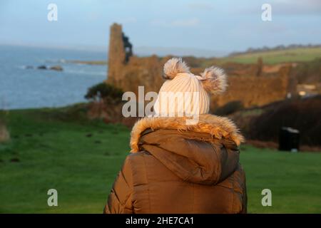 Dunure, Ayrshire, Schottland, Großbritannien. Eine Frau am Wintertag sitzt mit dem Rücken zur Kamera und schaut auf Dunure Castle Stockfoto