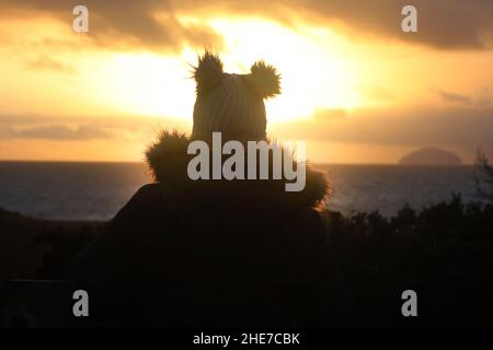 Dunure, Ayrshire, Schottland, Großbritannien. Eine Frau am Wintertag sitzt mit dem Rücken zur Kamera und schaut den Sonnenuntergang an der Ayrshire Coast an, mit einem warmen Bommel-Hut Stockfoto